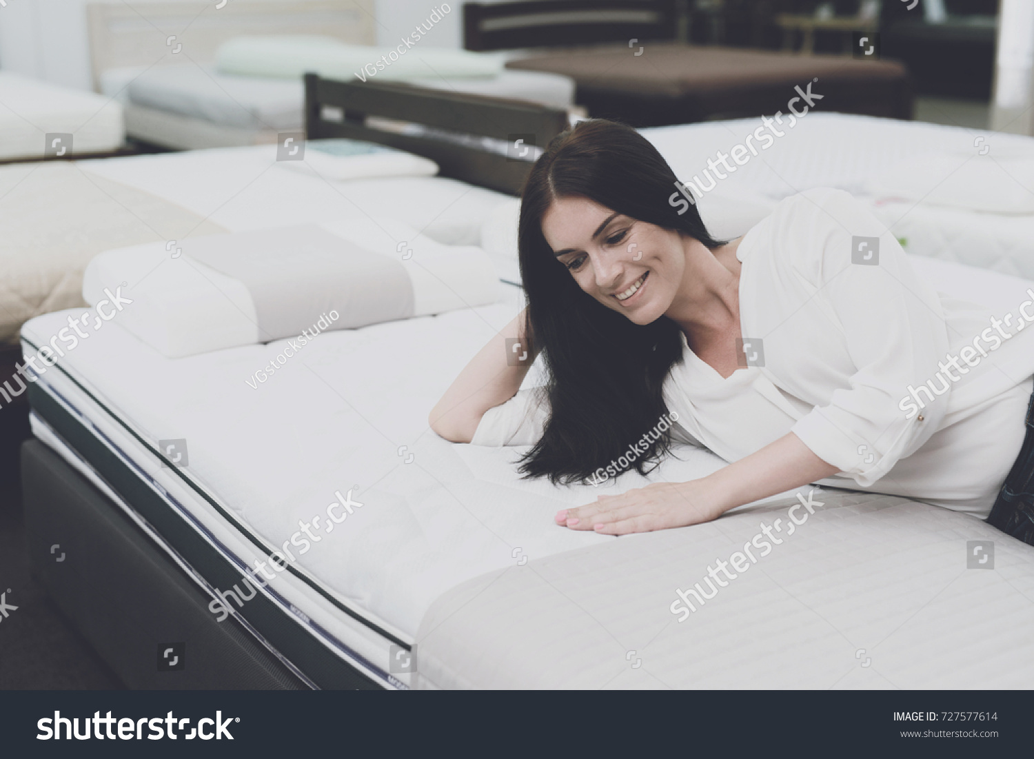 A woman in a white shirt and jeans in a mattress store. She examines the mattress she wants to buy. She lay down on the mattress and examines it. #727577614