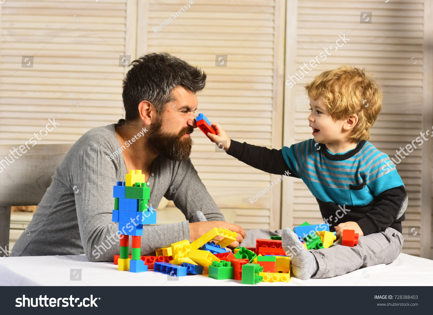 Family and childhood concept. Man with beard and boy play together on wooden wall background. Dad and kid build of plastic blocks. Father and son with happy faces create colorful robot with toy bricks #728388403