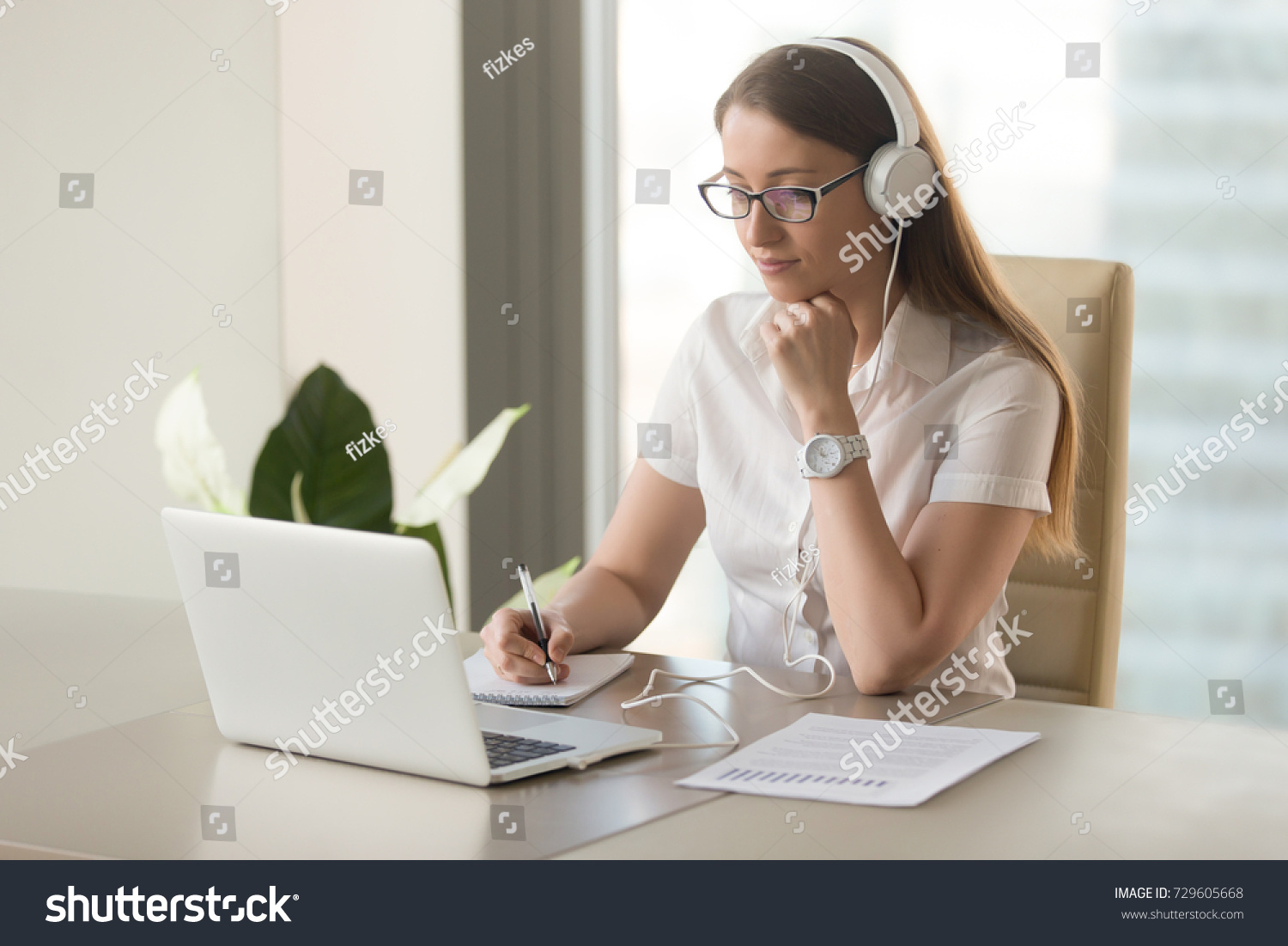 Focused attentive woman in headphones sits at desk with laptop, looks at screen, makes notes, learns foreign language in internet, online study course, self-education on web, consults client by video #729605668