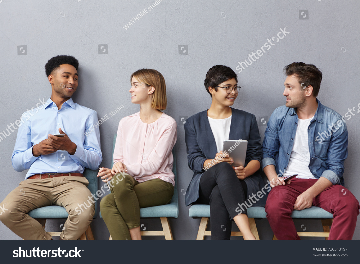 Horizontal portrait of people sit in queue, have pleasant conversation with each other, share ideas and life experience, isolated over grey concrete wall. Diverse group in row, speak and hold gadgets #730313197