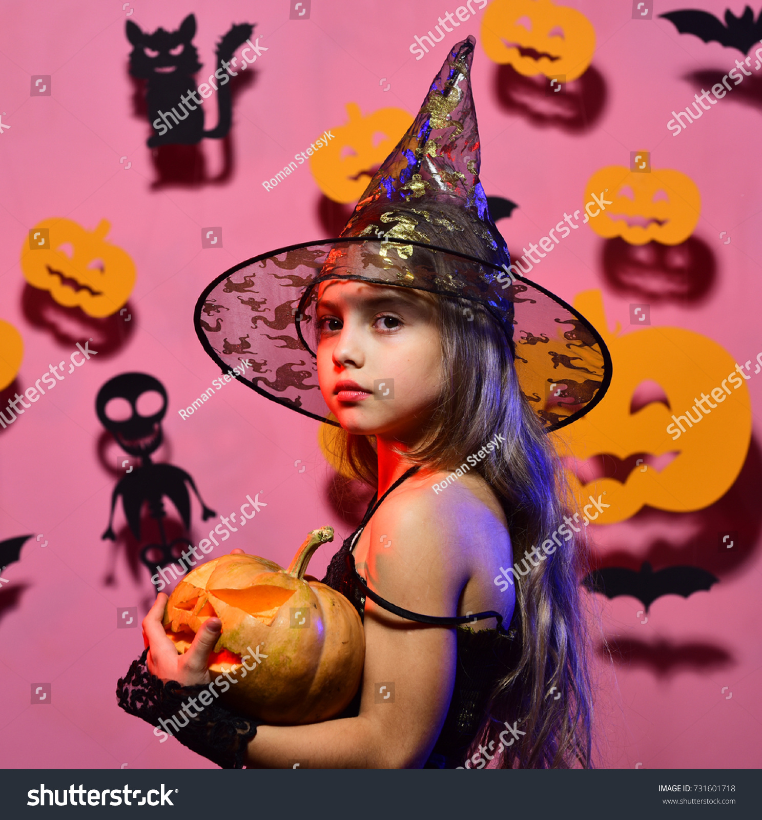 Little witch wearing black hat. Girl with calm face on pink background with bats and pumpkins decor. Halloween party and decorations concept. Kid in spooky witches costume holds carved pumpkin #731601718