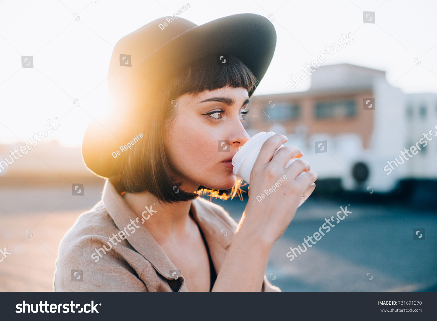 Attractive adorable woman with natural makeup sips on coffee in early morning on sunrise or sunset with light leaks, from to go cup, wears hipster fedora hat #731691370