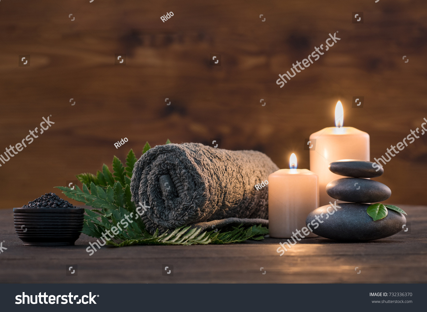 Towel on fern with candles and black hot stone on wooden background. Hot stone massage setting lit by candles. Massage therapy for one person with candle light. Beauty spa treatment and relax concept. #732336370