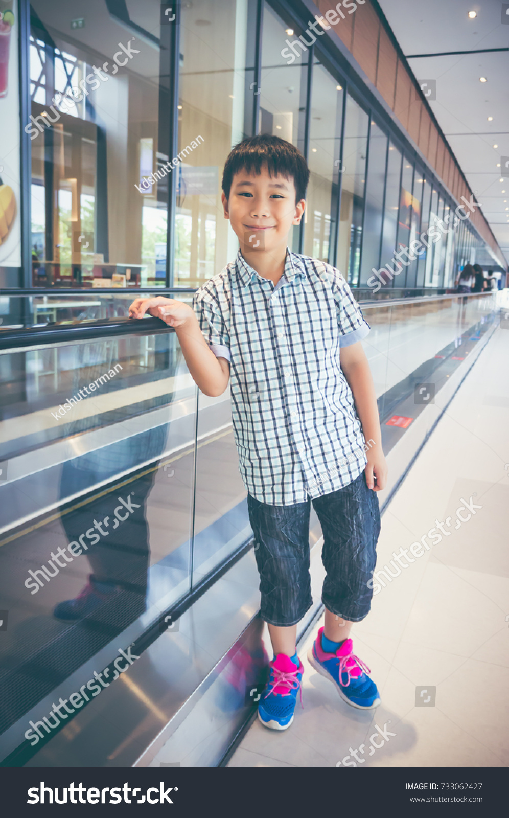 Happy asian child standing near electric speedwalk in modern airport. Handsome boy smiling and looking at camera. Travel on vacation. Vintage film filter effect. #733062427