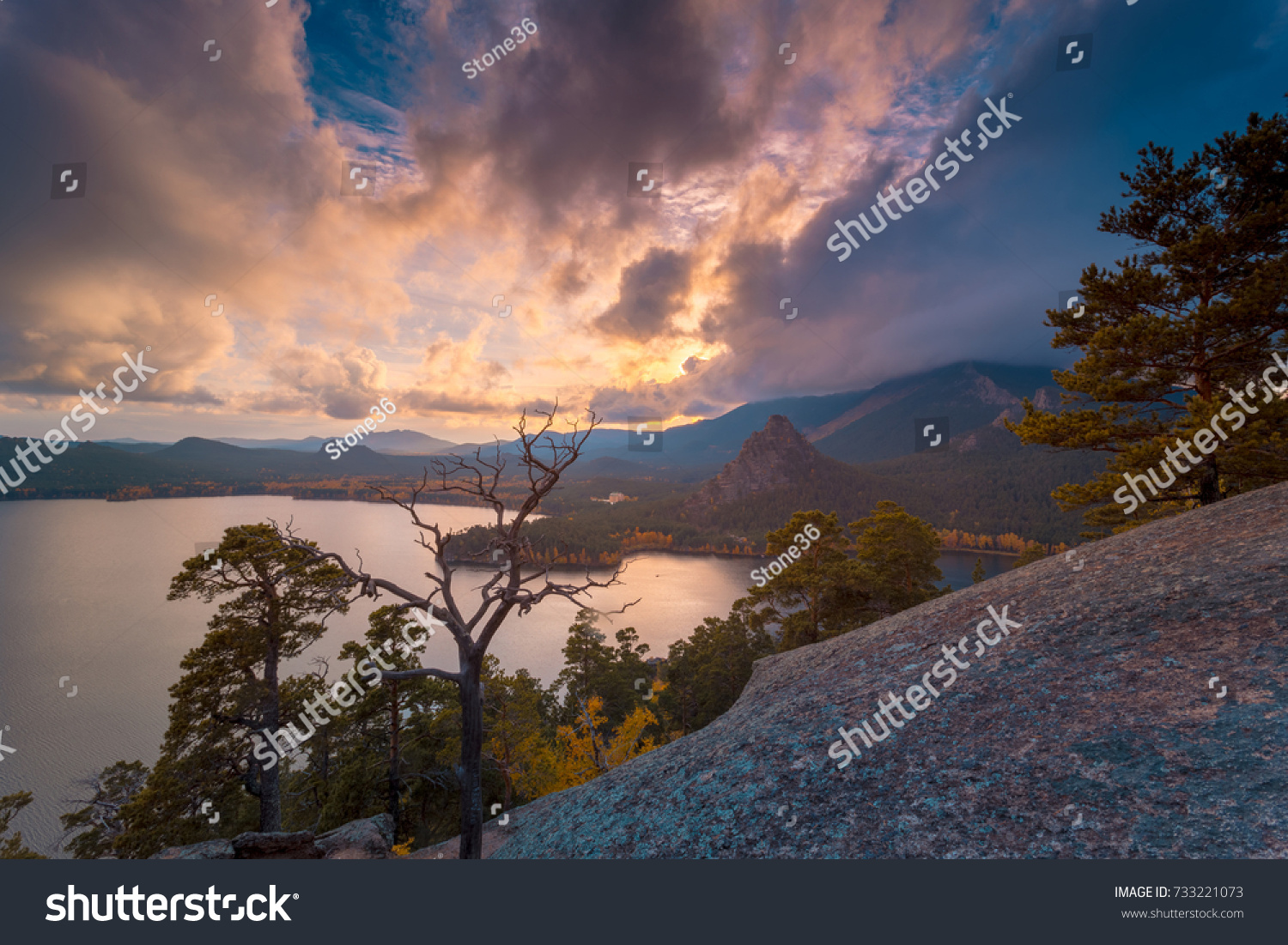 Beautiful sunset. Pine trees at sunset on a rocky mountains hill against dramatic clouds. Mountain lake on background. #733221073