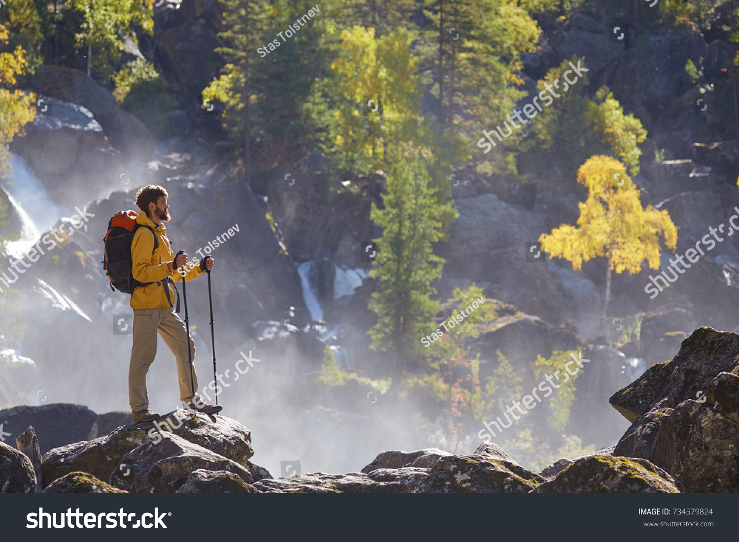 Hiker hiking with backpack looking at waterfall in park in beautiful autumn nature landscape. Portrait of male adult back standing outdoor. #734579824