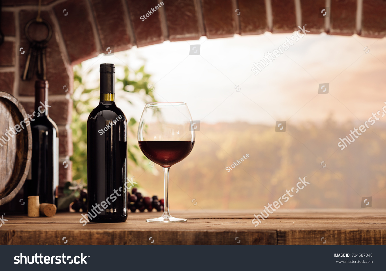 Red wine tasting in the wine cellar: wineglass and bottles next to the window and panoramic view of vineyards at sunset #734587048