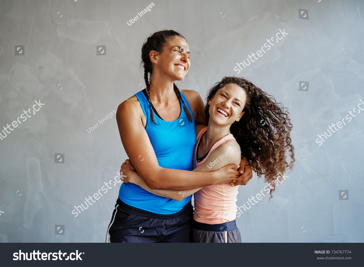 Two young women in sportswear laughing and standing arm in arm together in a gym #734767774