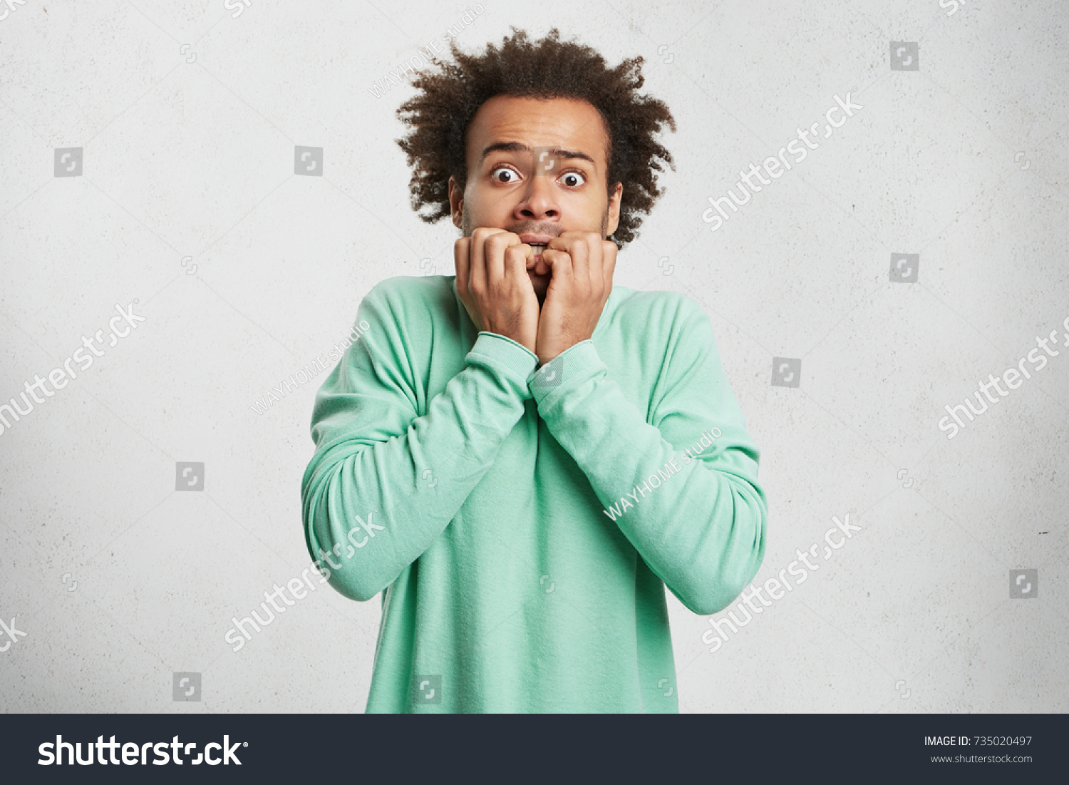 Portrait of young Afro American man with scared and anxious expression, bites finger nails, wears green sweater, being afraid of visiting doctor. Human facial expressions and negative emotions concept #735020497