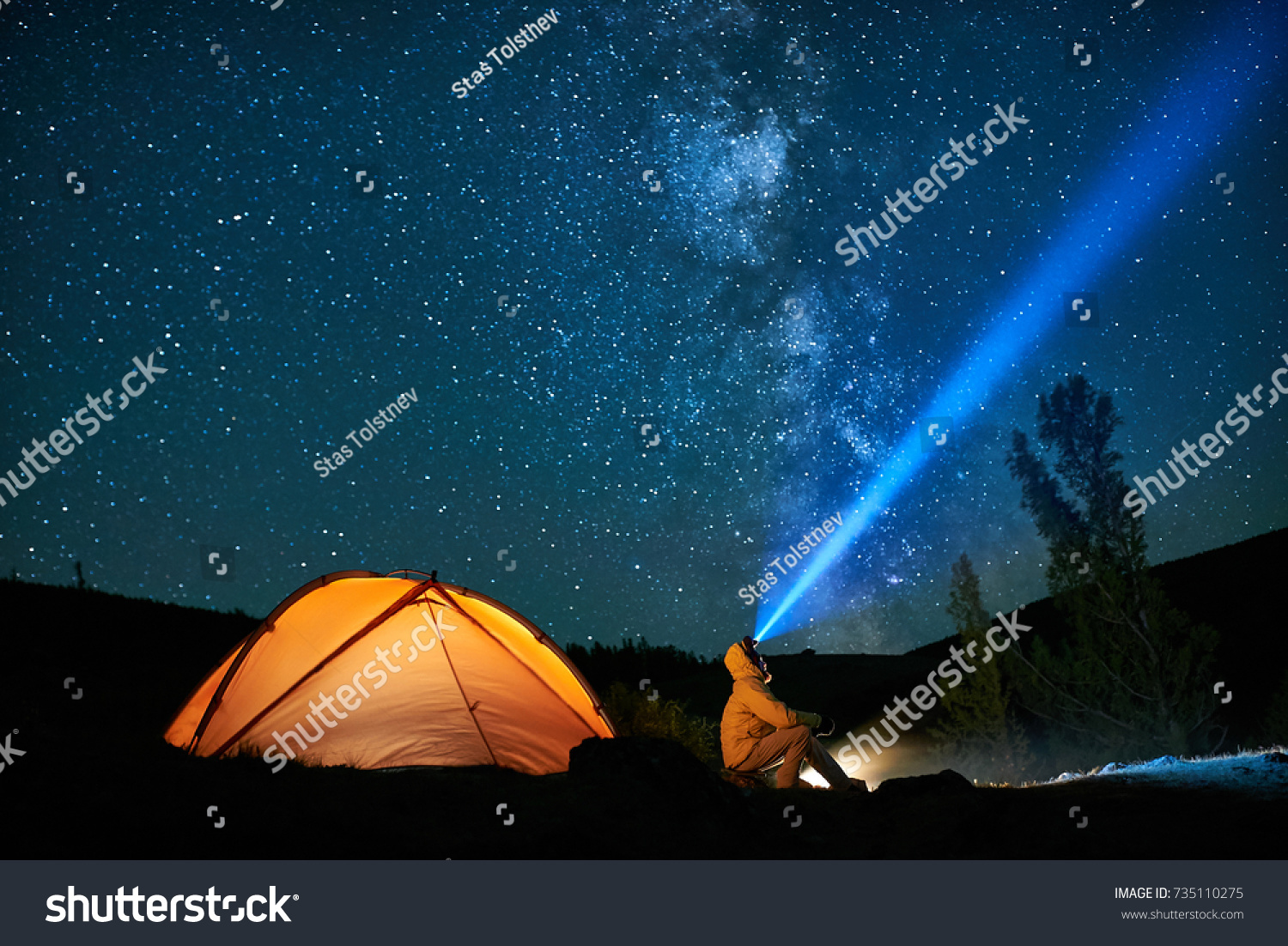 Man tourist with flashlight near his camp tent at night under a sky full of stars. Orange illuminated tent. #735110275