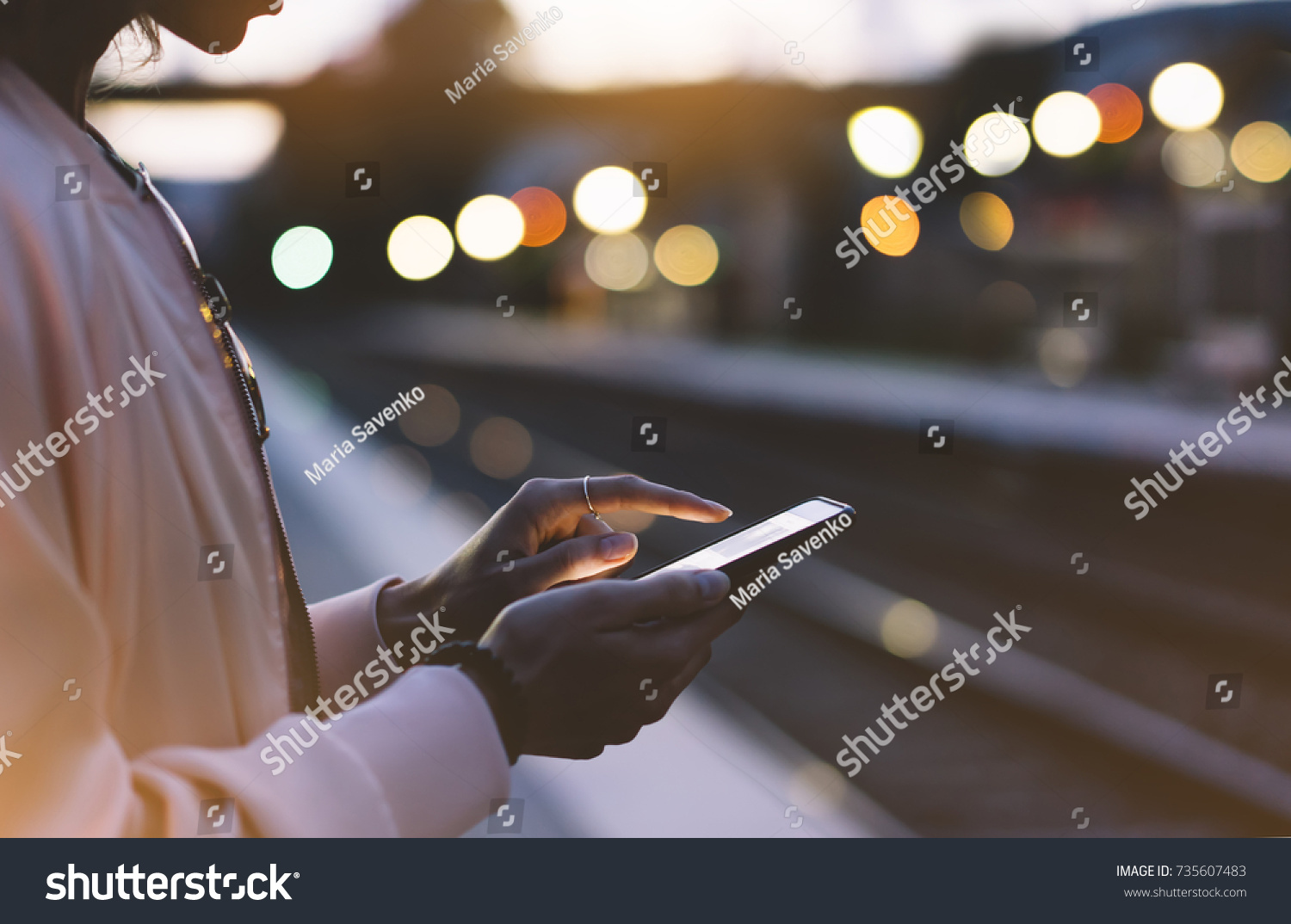 Woman waiting on station platform on background light train using smart phone in night. Tourist texting message and plan route of stop railway, railroad transport, booked. Enjoying travel concept #735607483
