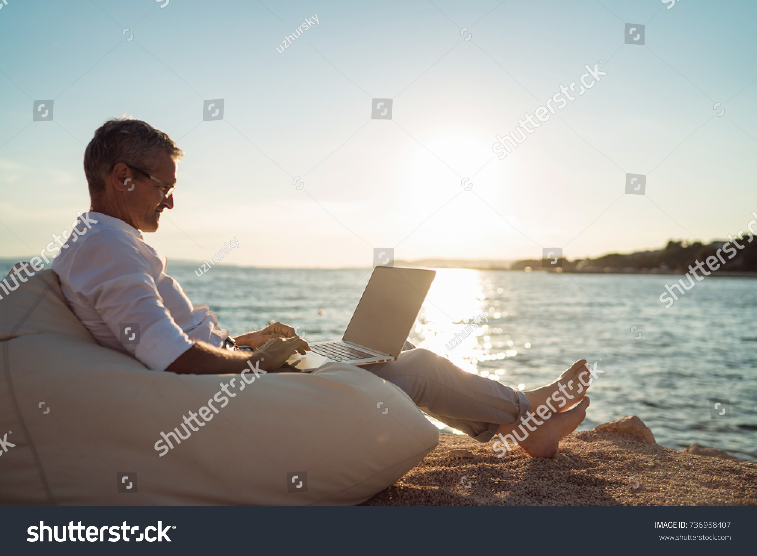 Senior man working on his laptop lying on deck chair on the beach during sunset #736958407