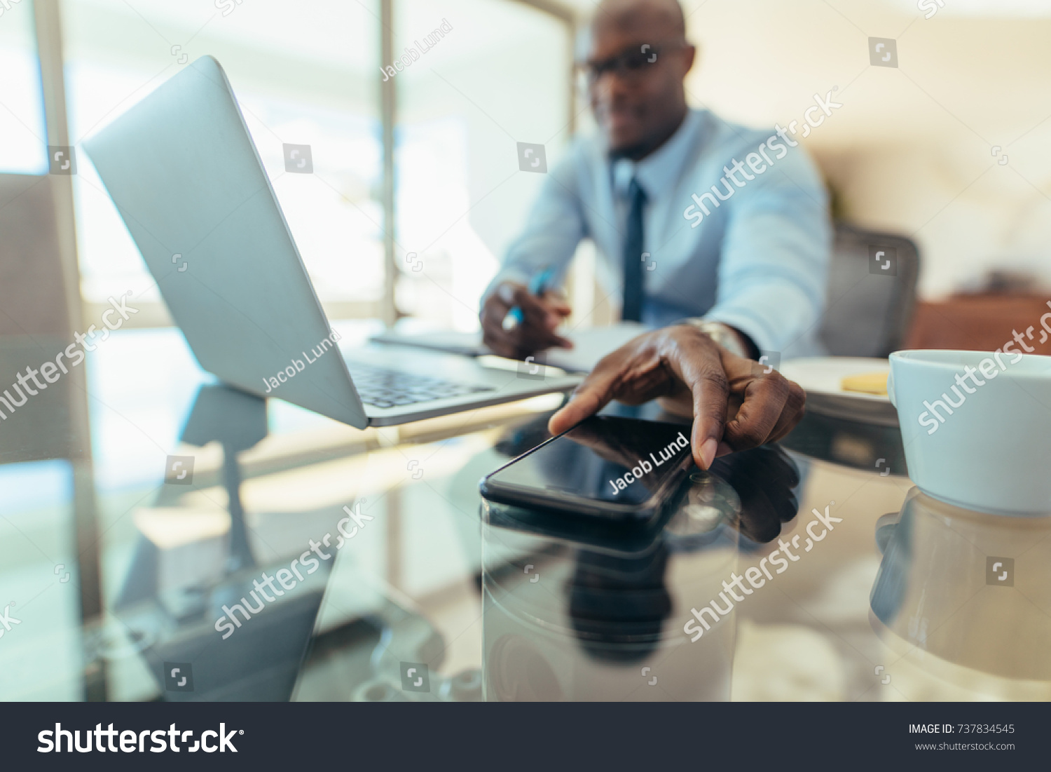 Man working on laptop computer sitting at his desk in office. Man reaching out to pick his mobile phone placed on table. #737834545