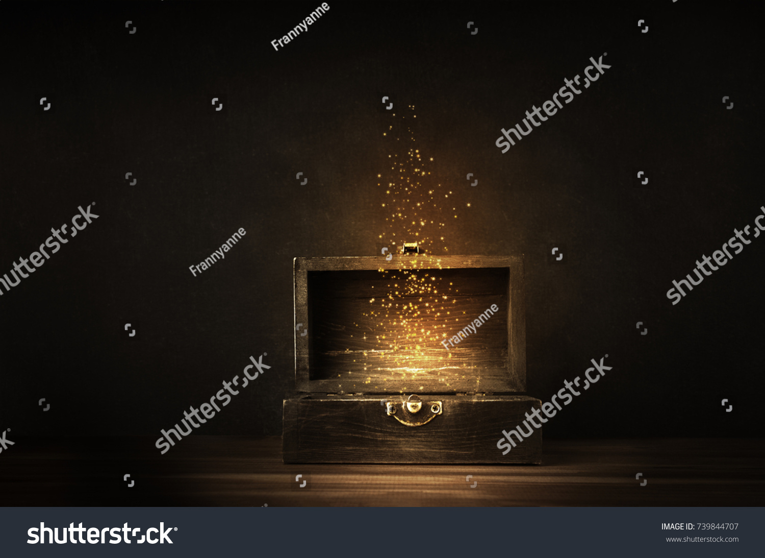 Glowing golden sparkles and stars rising from an old, opened wooden treasure chest. Darkly lit on a planked surface with black chalkboard background. #739844707