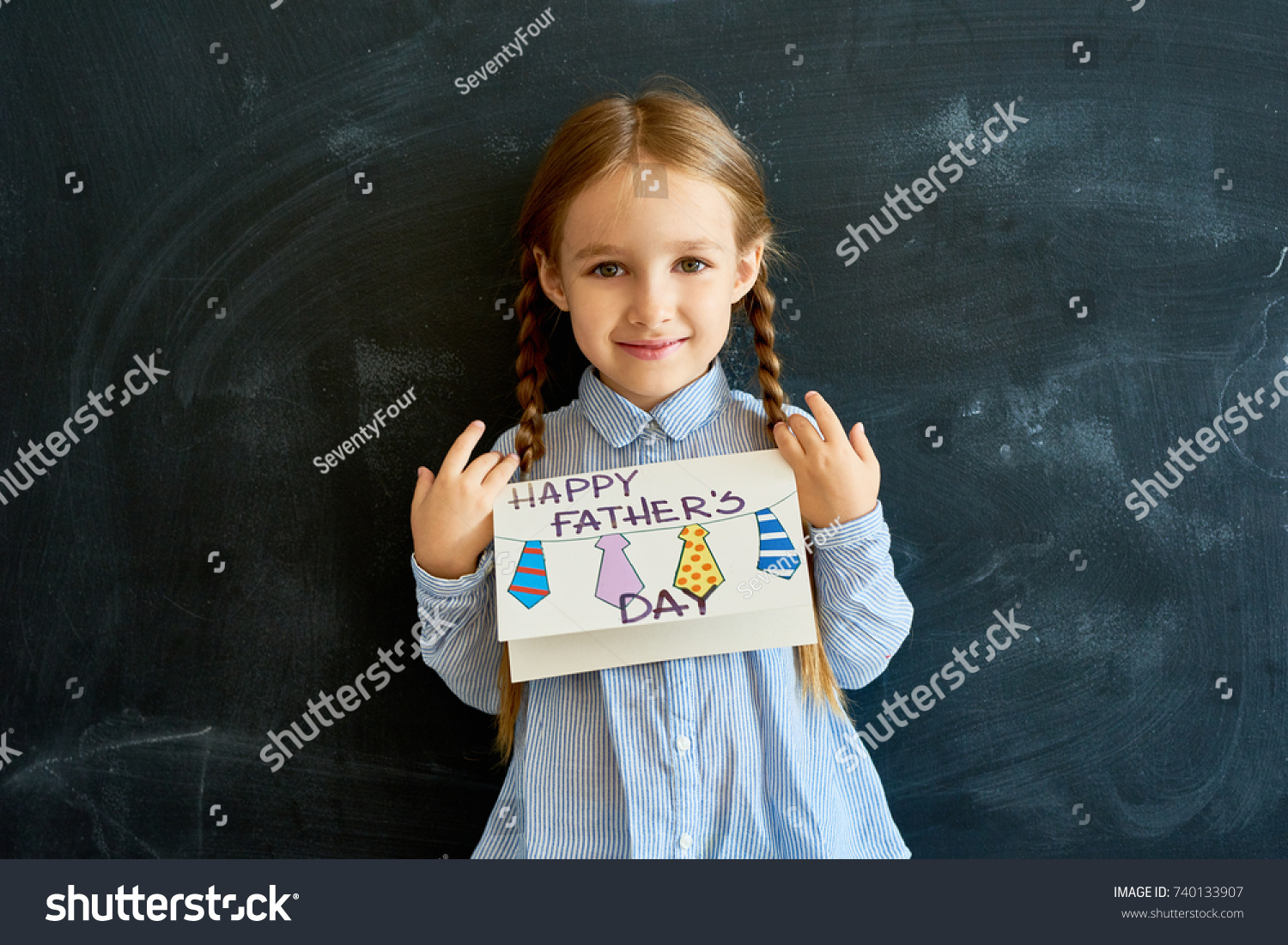 Portrait of happy little girl holding handmade  greeting card for Fathers Day posing against blackboard in school and looking at camera #740133907