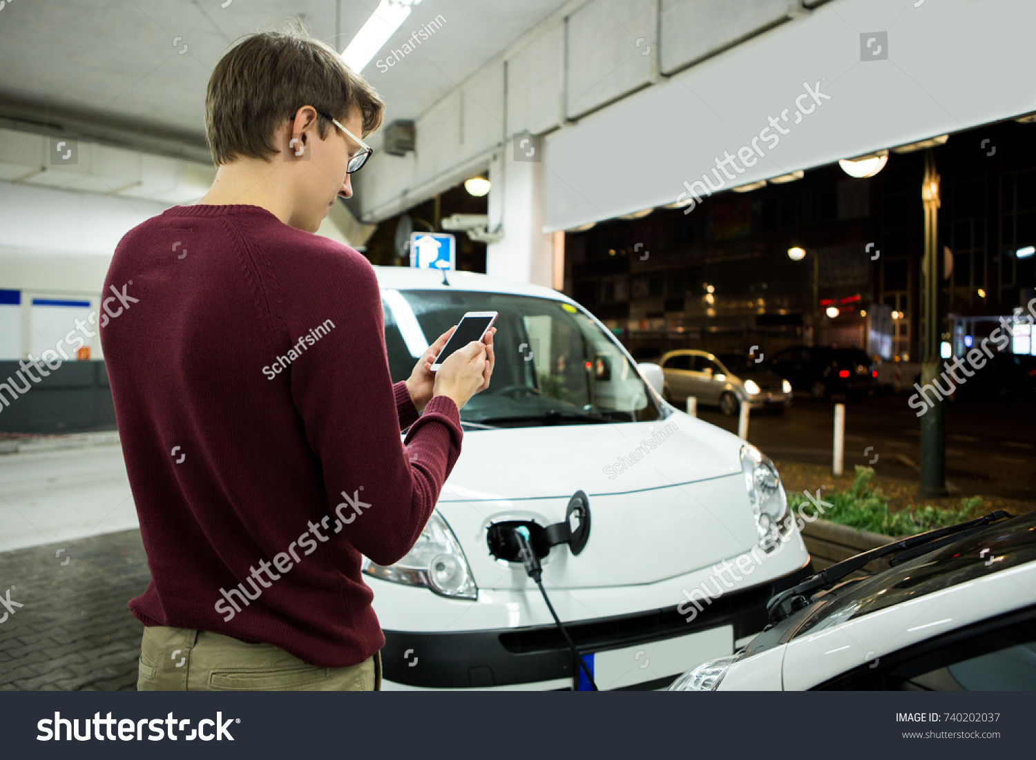 Man is holding a phone. His electric car charged at the charging station #740202037