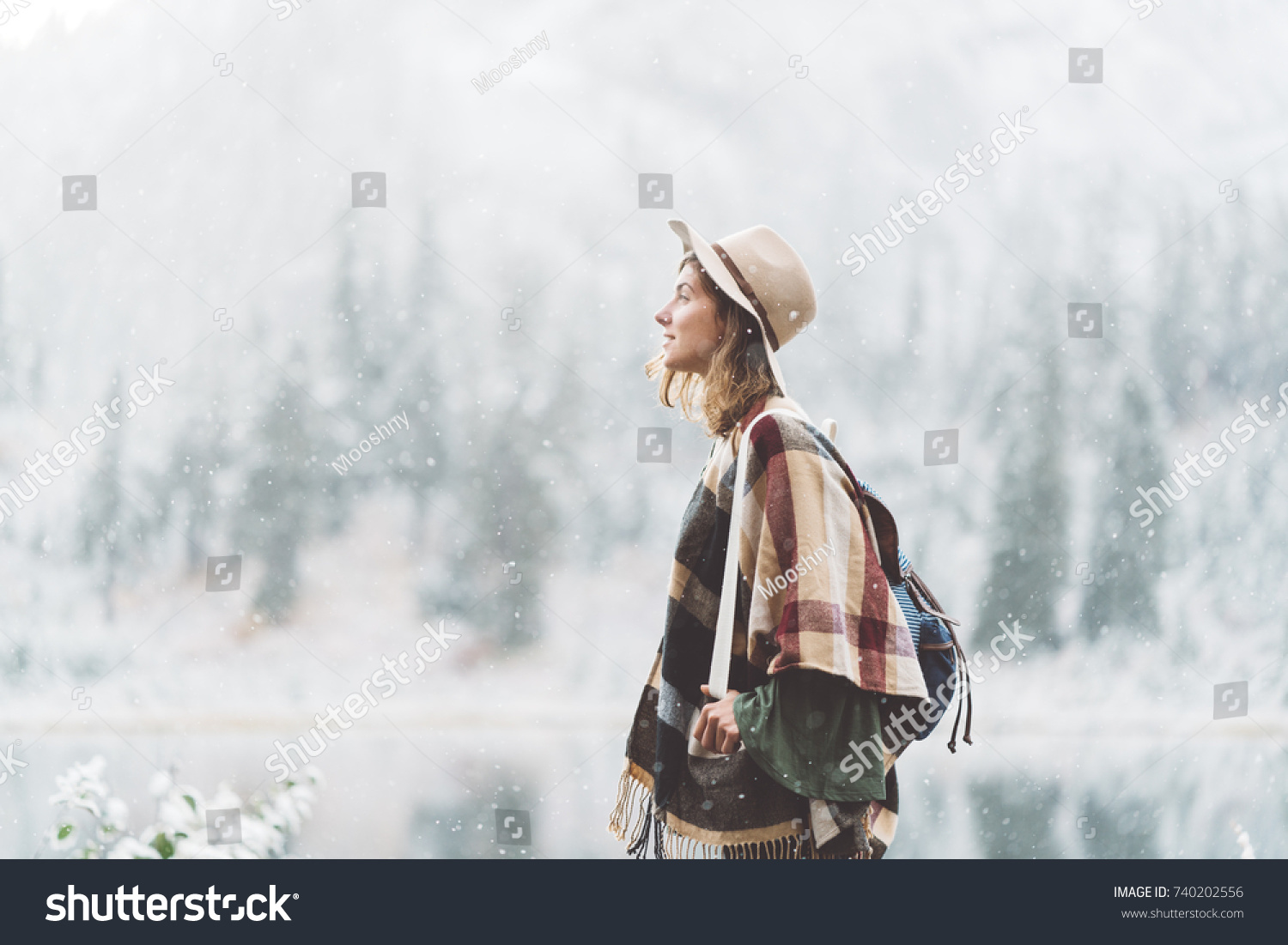 Handsome traveling woman enjoying wilderness in front of incredible mountain lake. Wearing hat, poncho and backpack. Winter is coming, first snowfall. Wanderlust and boho style #740202556