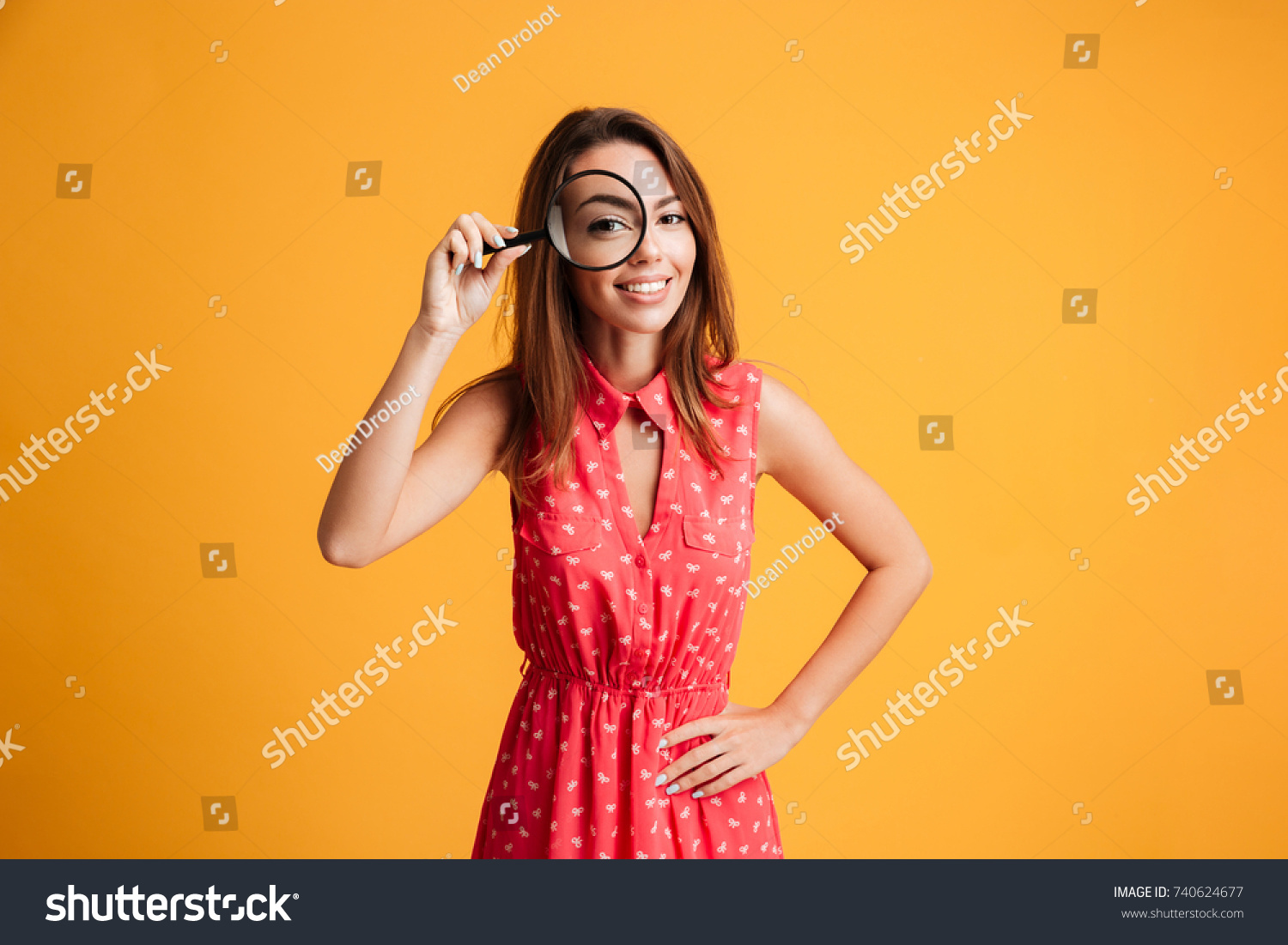 Close-up portrait of cheerful pretty woman in red dress looking at camera through magnifying glass, isolated over yellow background #740624677