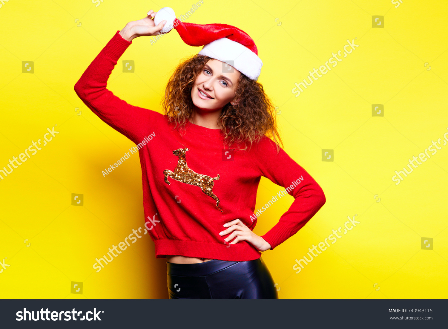 Santa woman in christmas hat wearing red sweater with deer smiling having fun laughing rejoices touches touches on yellow background in the studio. The concept of the holiday of Christmas and New Year #740943115