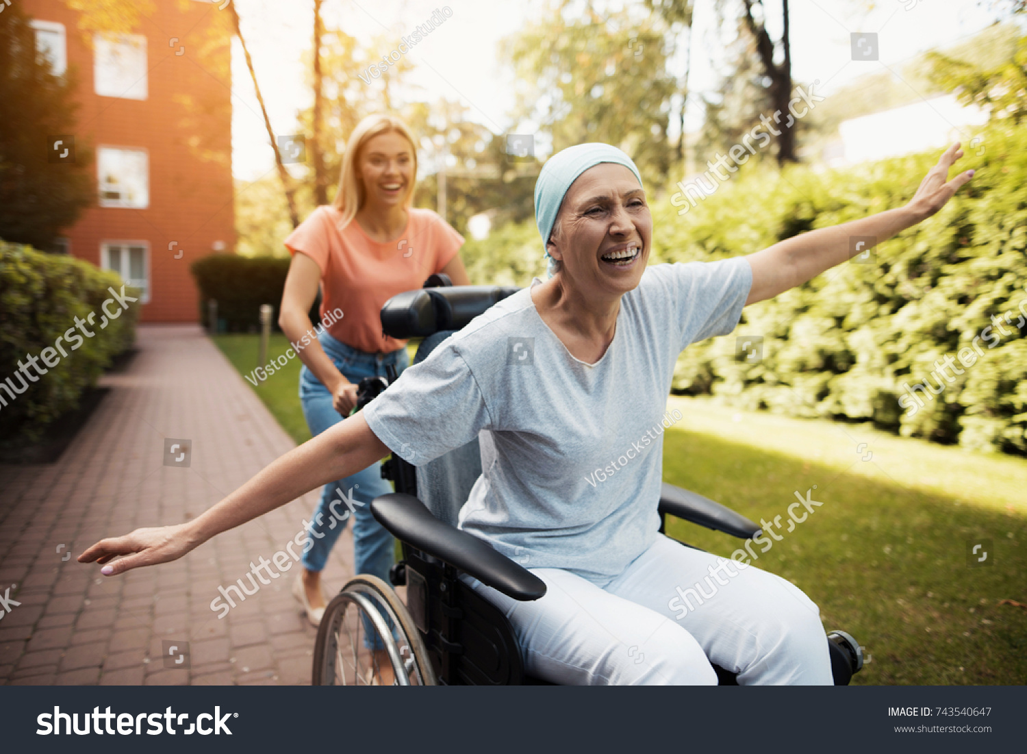 A woman with cancer is sitting in a wheelchair. She walks on the street with her daughter and they fool around. They are fun and they laugh. They walk in the courtyard of the clinic. #743540647