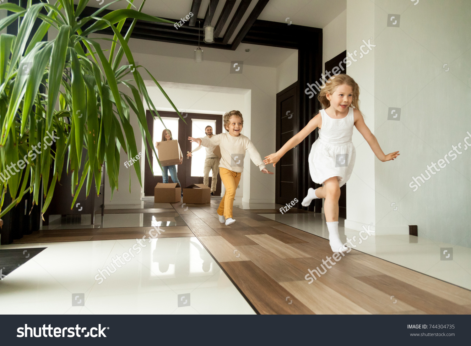 Happy young family with cardboard boxes in new home at moving day concept, excited children running into big modern own house hallway, parents with belongings at background, mortgage loan, relocation  #744304735