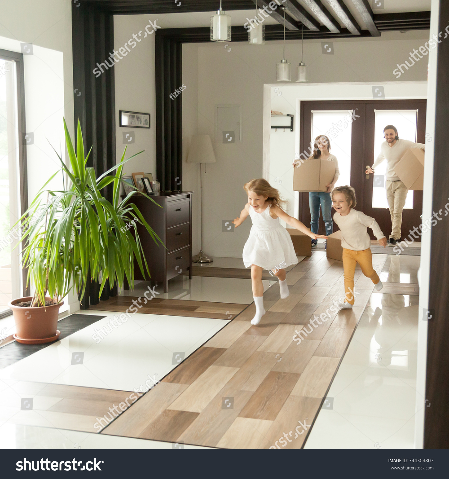Playful happy kids running into new big own beautiful house, family moving in day concept, excited children exploring home interior having fun together, parents holding cardboard boxes at background  #744304807