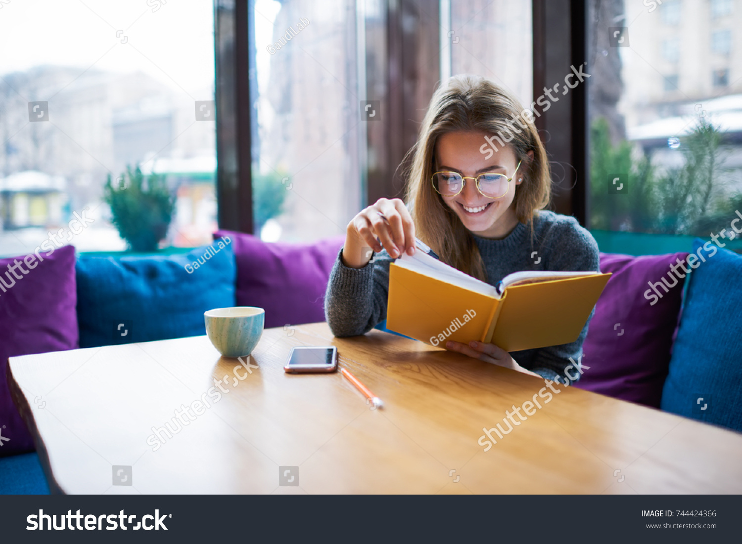 Smart student in optical eyeglasses enjoying favourite book in stylish coffee shop interior.Smiling hipster girl in spectacles reading funny stories sitting in coworking space with cup of cappuccino #744424366