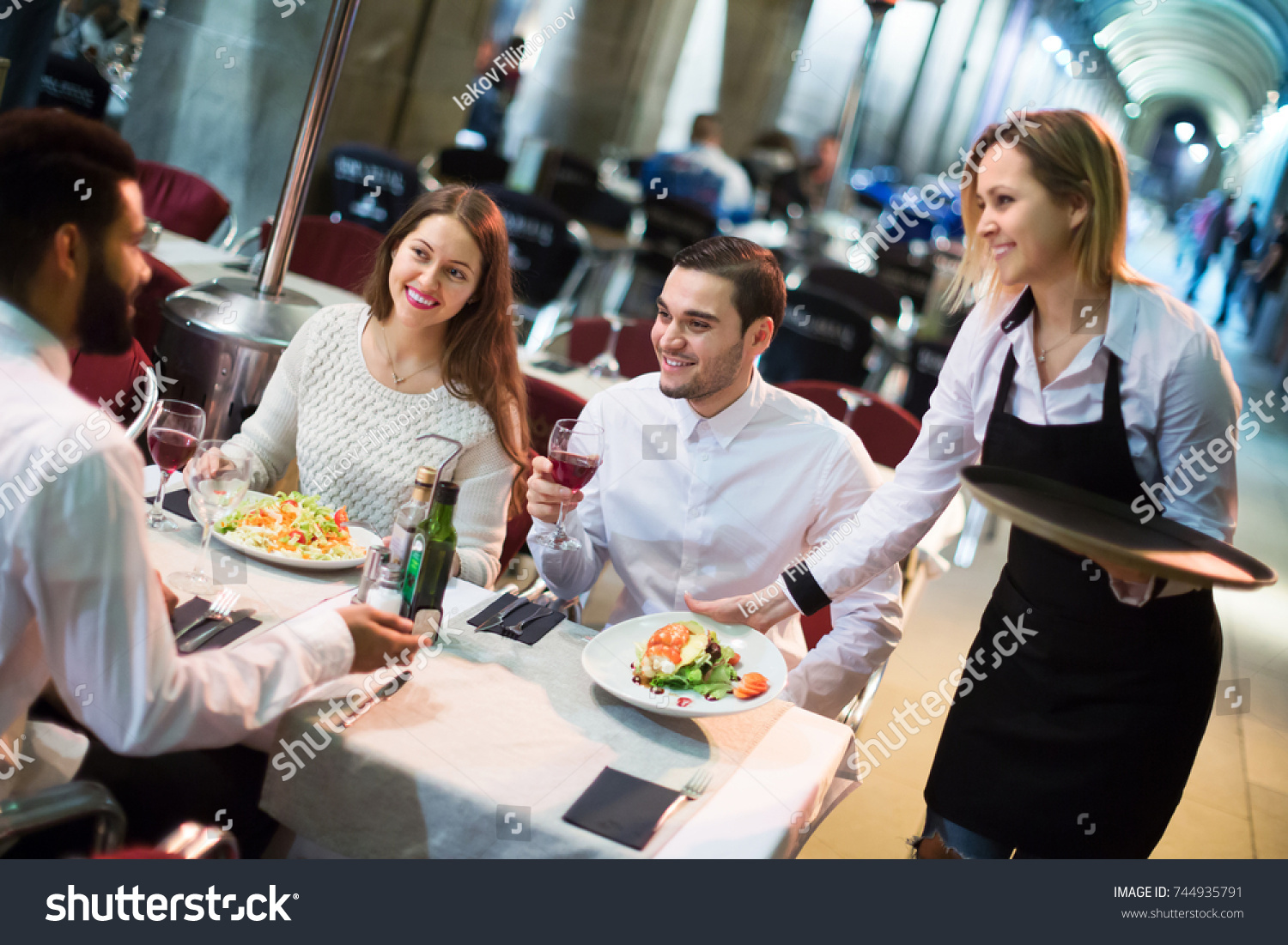 Portrait of smiling young friends in outdoors restaurant and smiling waitress #744935791