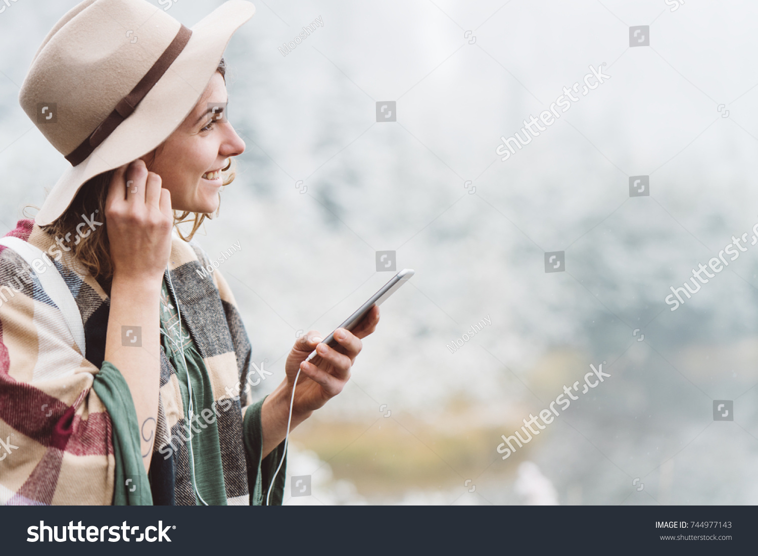 Smiling handsome woman listening music with her mobile phone in first snow. Traveling among stunning winter landscape. Vacations in mountain wilderness. Wanderlust and boho style #744977143