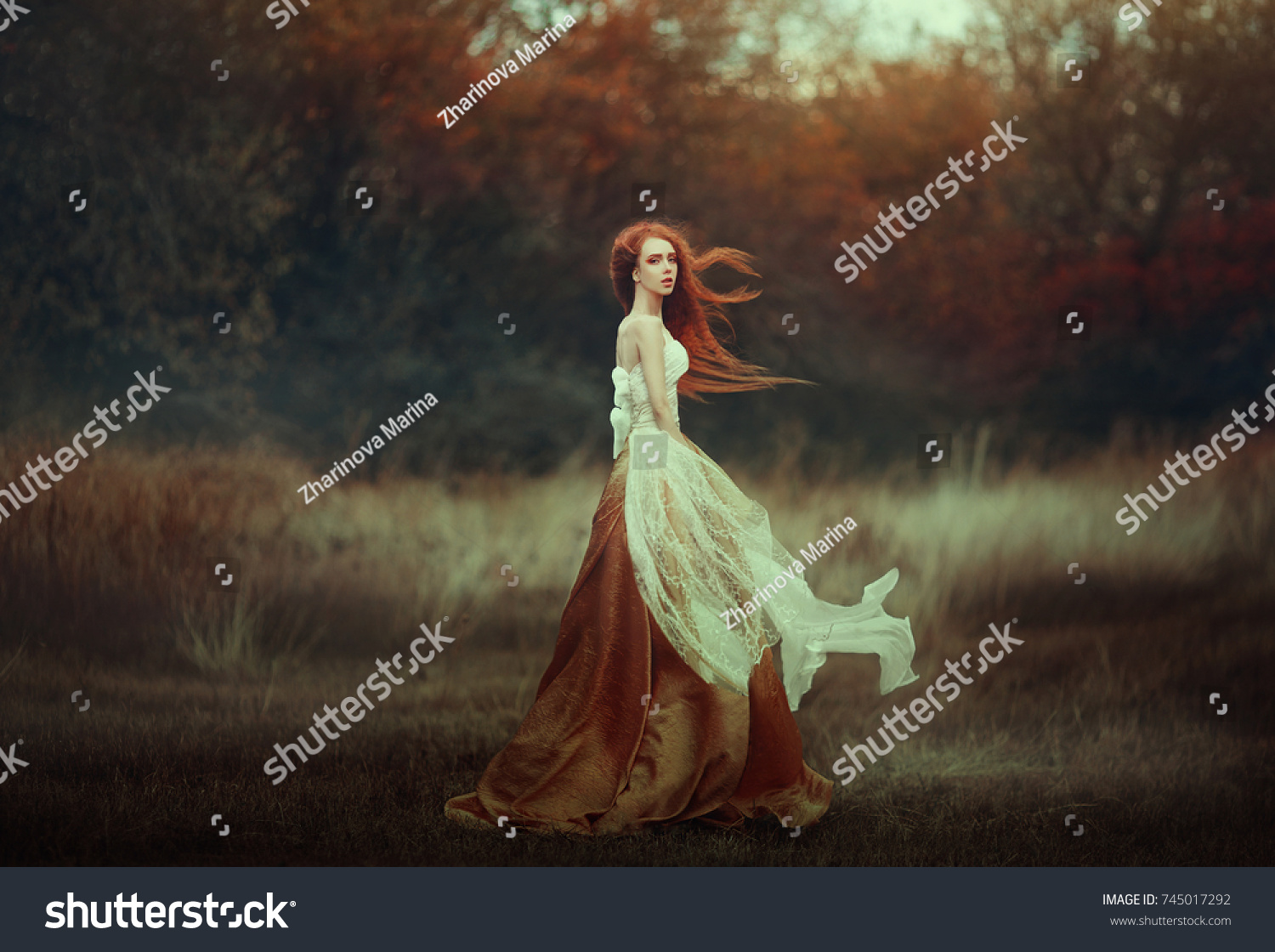 Beautiful young woman with very long red hair in a golden medieval dress walking through the autumn forest. Long red hair develops in the wind. #745017292