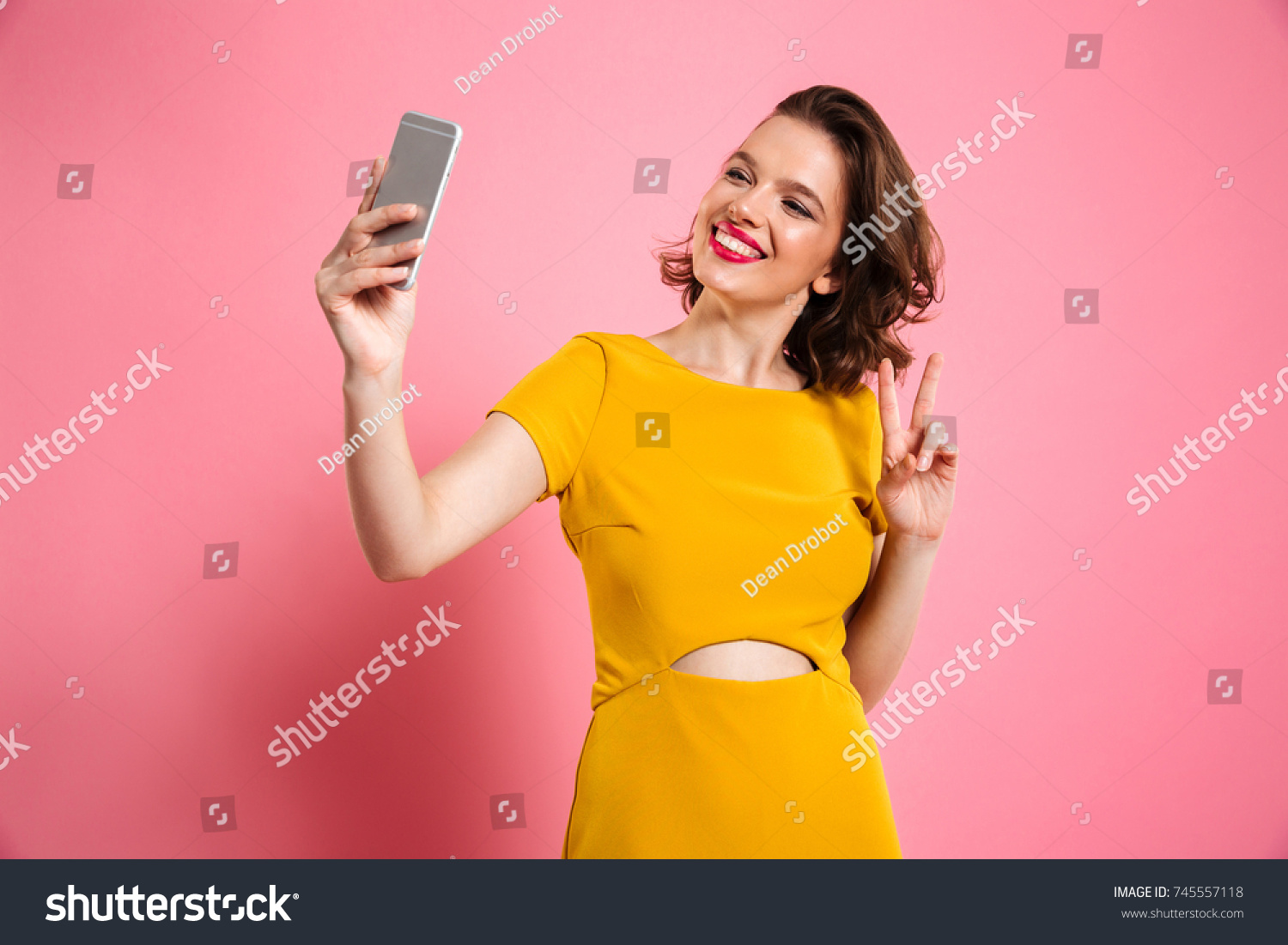 Cute pretty girl with bright makeup showing peace gesture while taking selfie on mobile phone, isolated over pink background #745557118