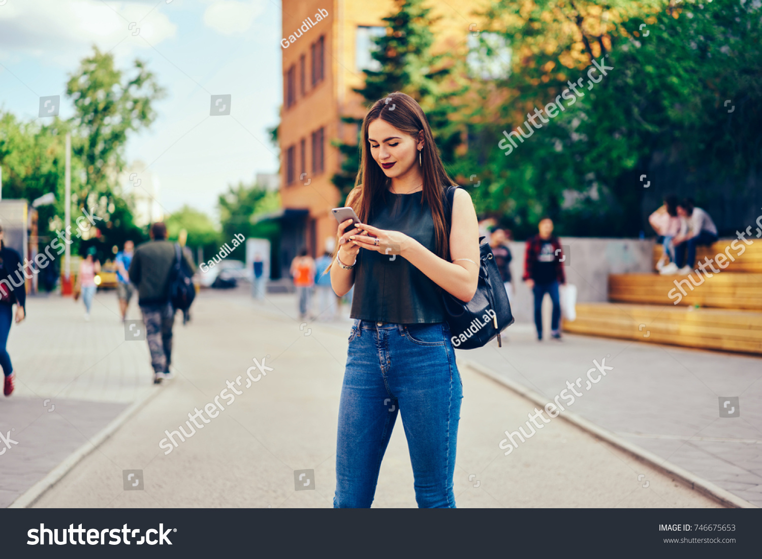 Beautiful young woman dressed in trendy clothes checking notification on smartphone while having summer walk outdoors.Pretty pensive hipster girl strolling at street and sending messages on cellular #746675653