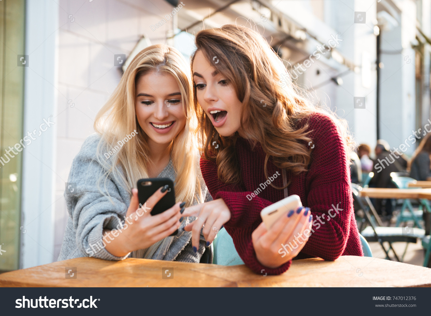Two excited young girls using mobile phones while sitting at the cafe outdoors and pointing finger #747012376