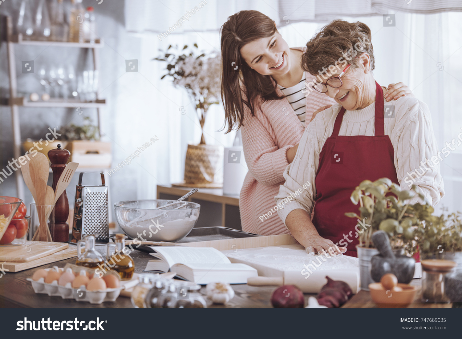 Smiling woman massaging grandmother's shoulder while making cake in the kitchen #747689035