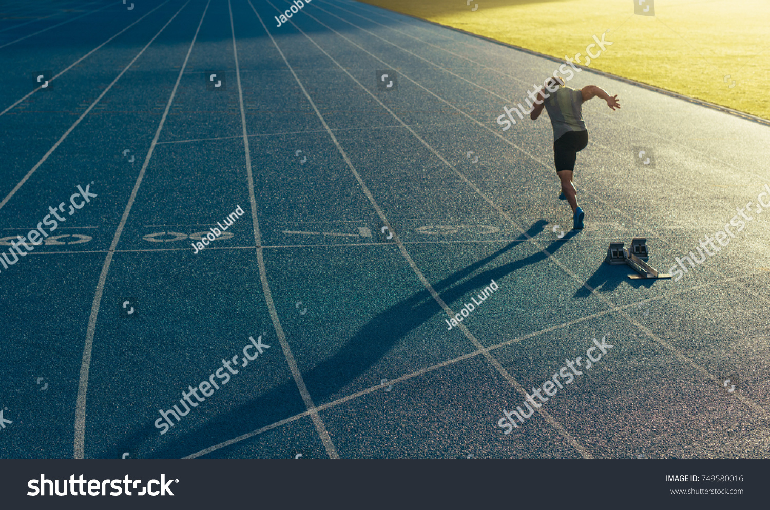 Athlete running on an all-weather running track alone. Runner sprinting on a blue rubberized running track starting off using a starting block. #749580016