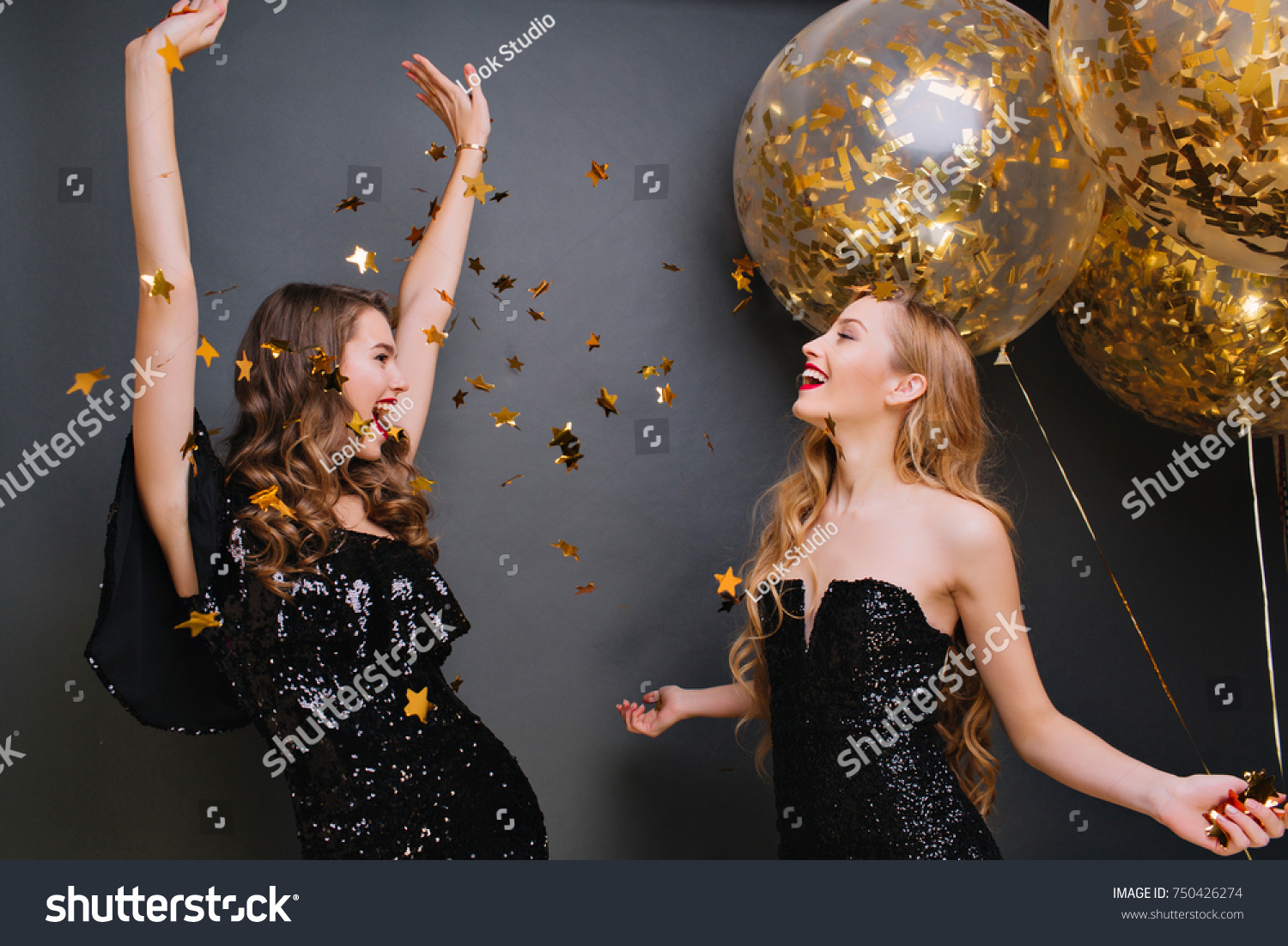 Inspired curly pale woman singing with hands up on dark background. Romantic blonde girl in black outfit holding party balloons and looking at friend which dancing under confetti. #750426274