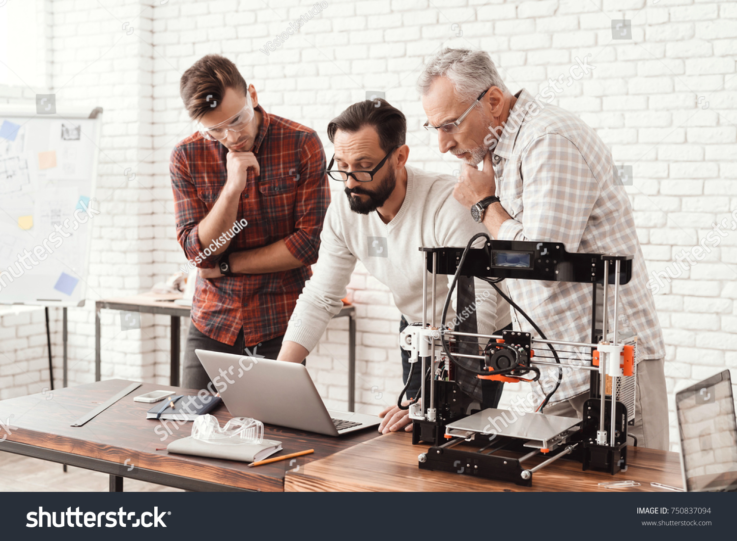 Three men are working on preparing a 3d printer for printing. One of them explains the rest of the subtlety to the print. A young and elderly man is looking at the laptop monitor screen. #750837094
