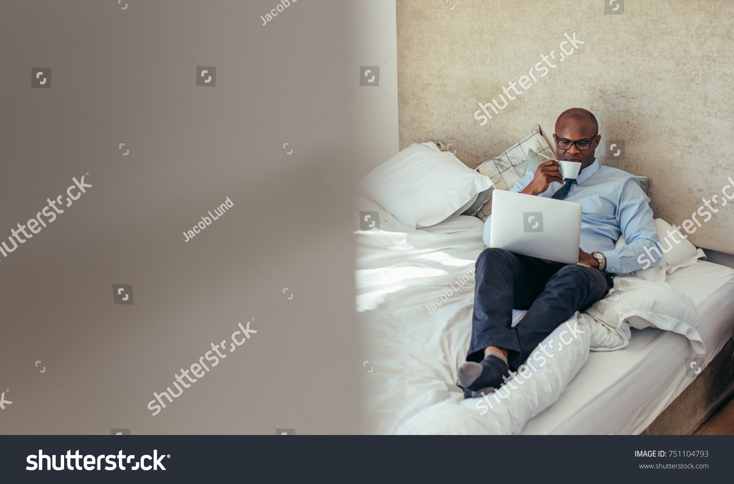 Man in formal clothes working on laptop while lying in bed. Businessman working on laptop computer while drinking coffee in his hotel room. #751104793