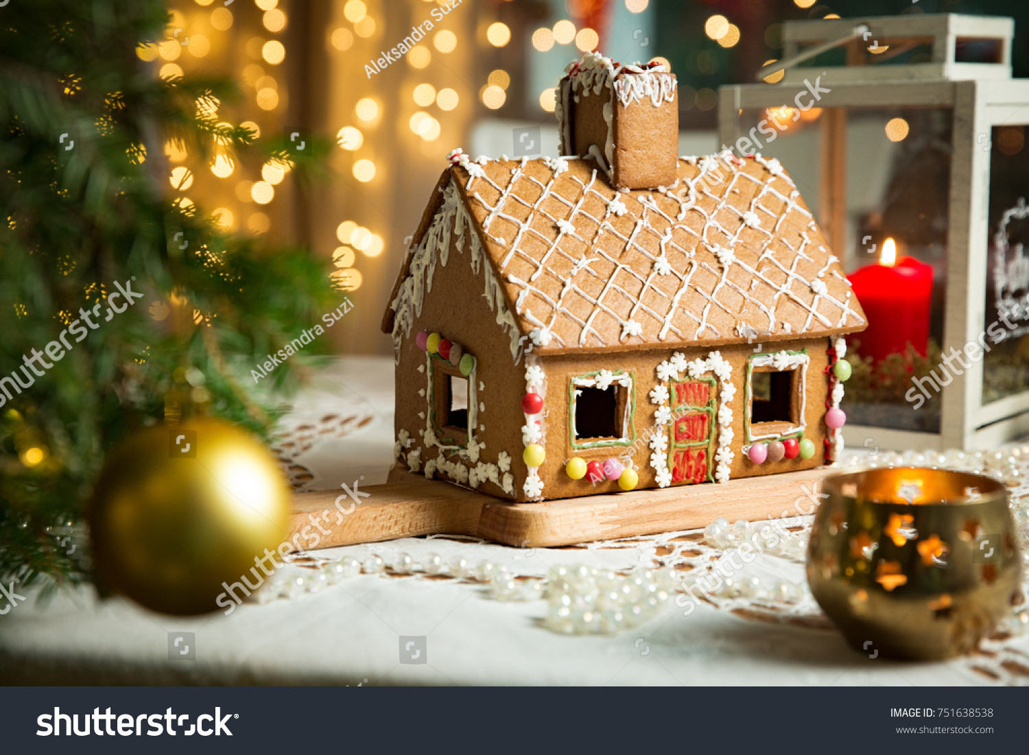 Little gingerbread house with glaze standing on table with tablecloth and decorations, candles and lanterns. Living room with lights and Christmas tree. Holiday mood #751638538
