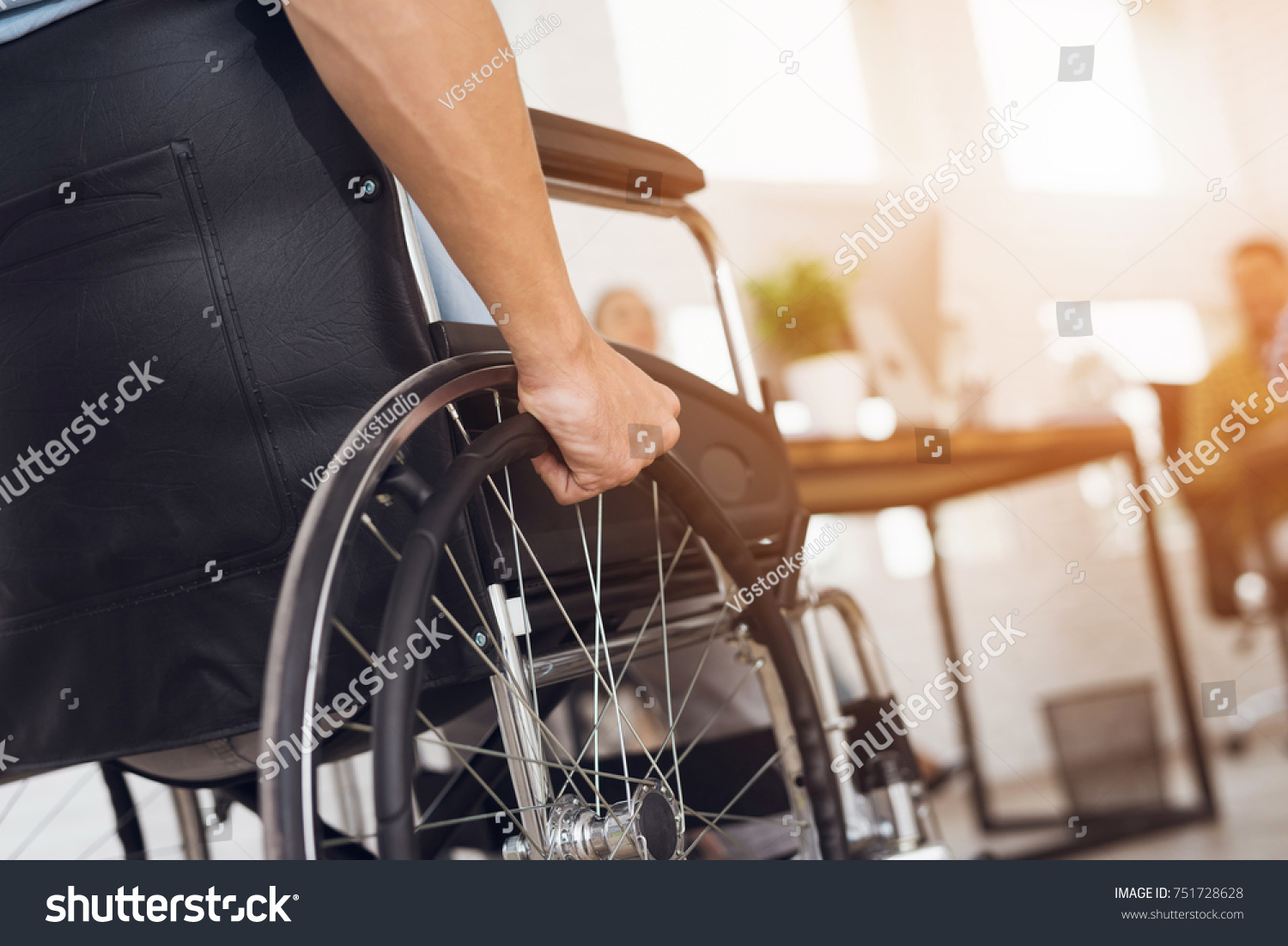 A disabled man is sitting in a wheelchair. He holds his hands on the wheel. Nearby are his colleagues #751728628