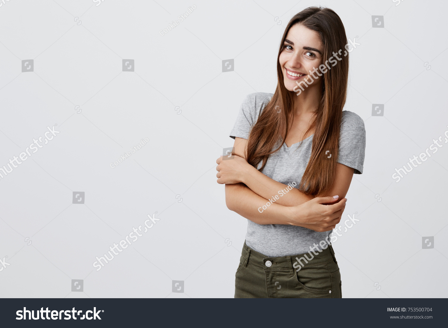 Cheerful young beautiful brunette caucasian student girl with long hair in casual outfit smiling brightly, holding hands together, posing for university graduation photo in light studio. Copy space #753500704