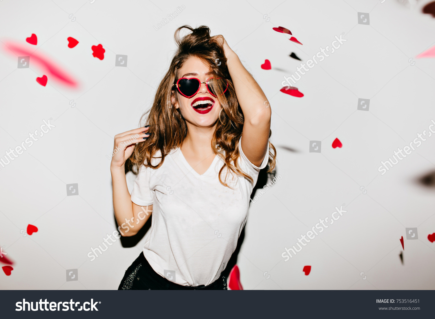 Indoor portrait of amazing caucasian female model in trendy t-shirt touching her long shiny hair. Laughing refined woman in sunglasses having fun with red confetti. #753516451