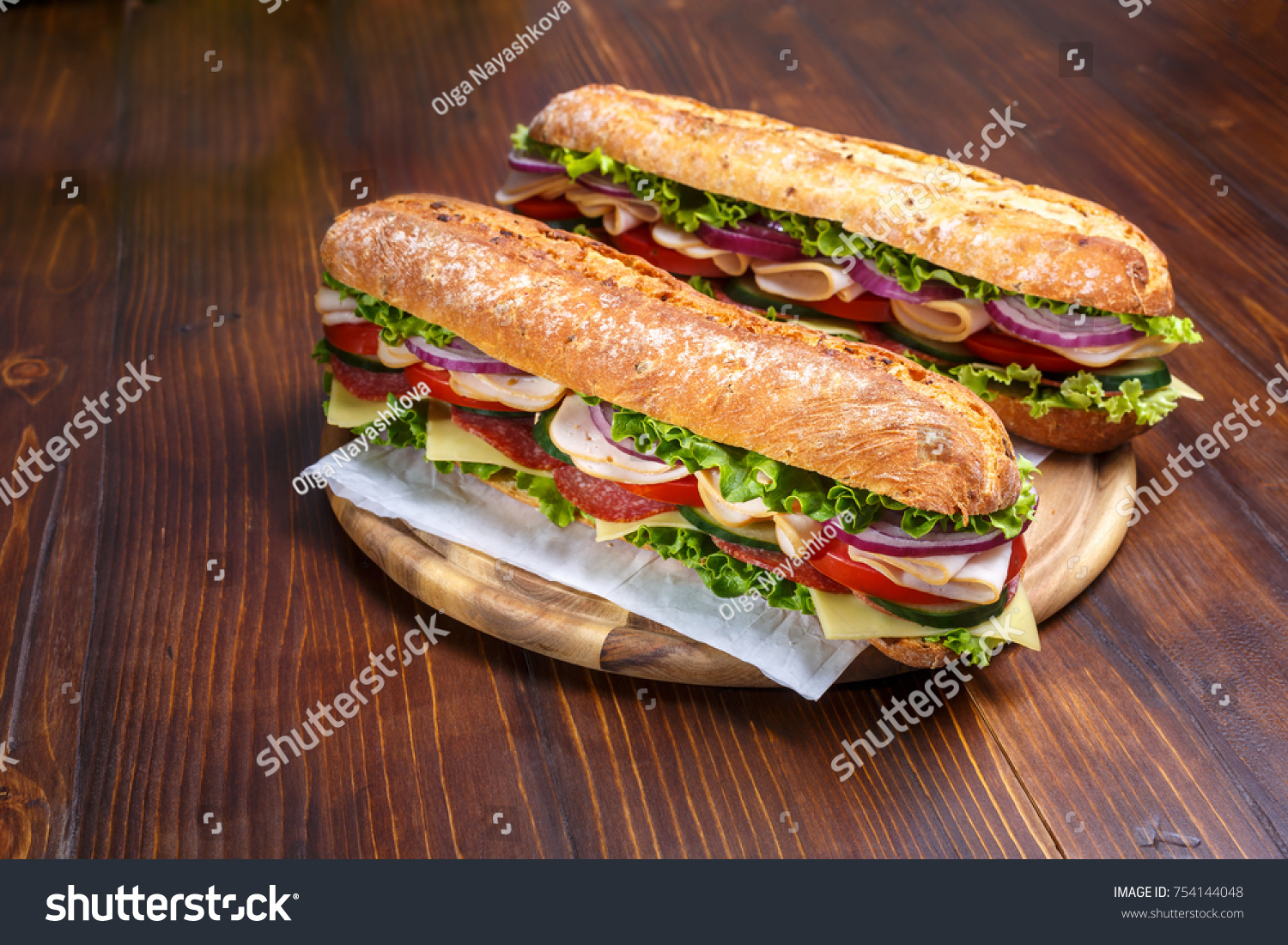 Two baguette sandwiches with salami, turkey breast, cheese, lettuce, tomatoes and onion on a cutting board. Subway sandwiches on a dark background.
 #754144048