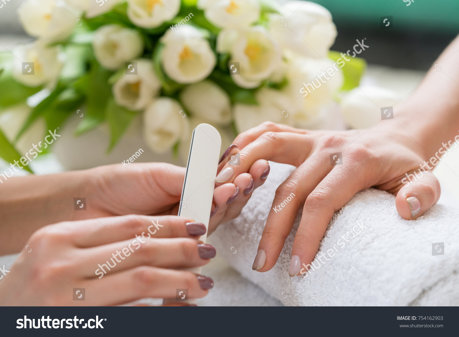 Close-up of the hands of a qualified manicurist filing the nails of a young woman with a white buffer in a trendy nail salon #754162903