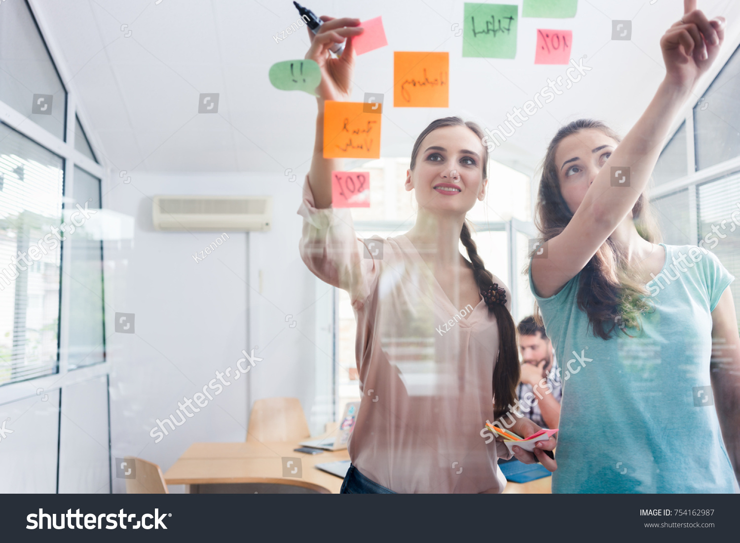 Two proficient female co-workers posting sticky notes in the interior of a shared office space for task prioritization and better time management #754162987