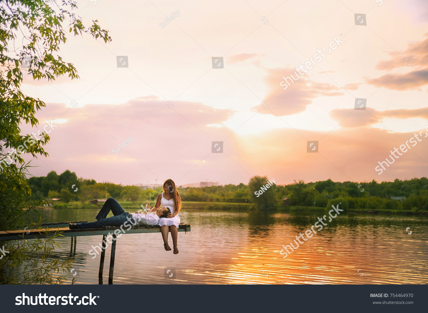 
Beautiful bright fairy walk of a loving couple in the summer at sunset. A guy in a white shirt and jeans with a girl in a sarafan embraces, dancing, sitting on the pier. Pond in the background. Place #754464970