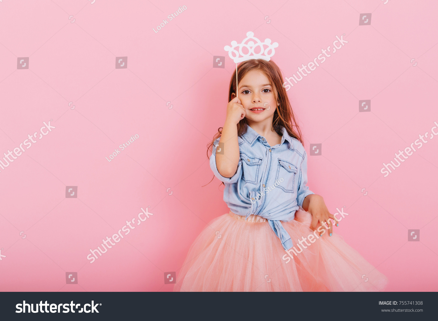 Pretty sweet little girl with long brunette hair in tulle skirt holding white crown on head isolated on pink background. Beautiful joyful child expressing true emotions. Place for text #755741308