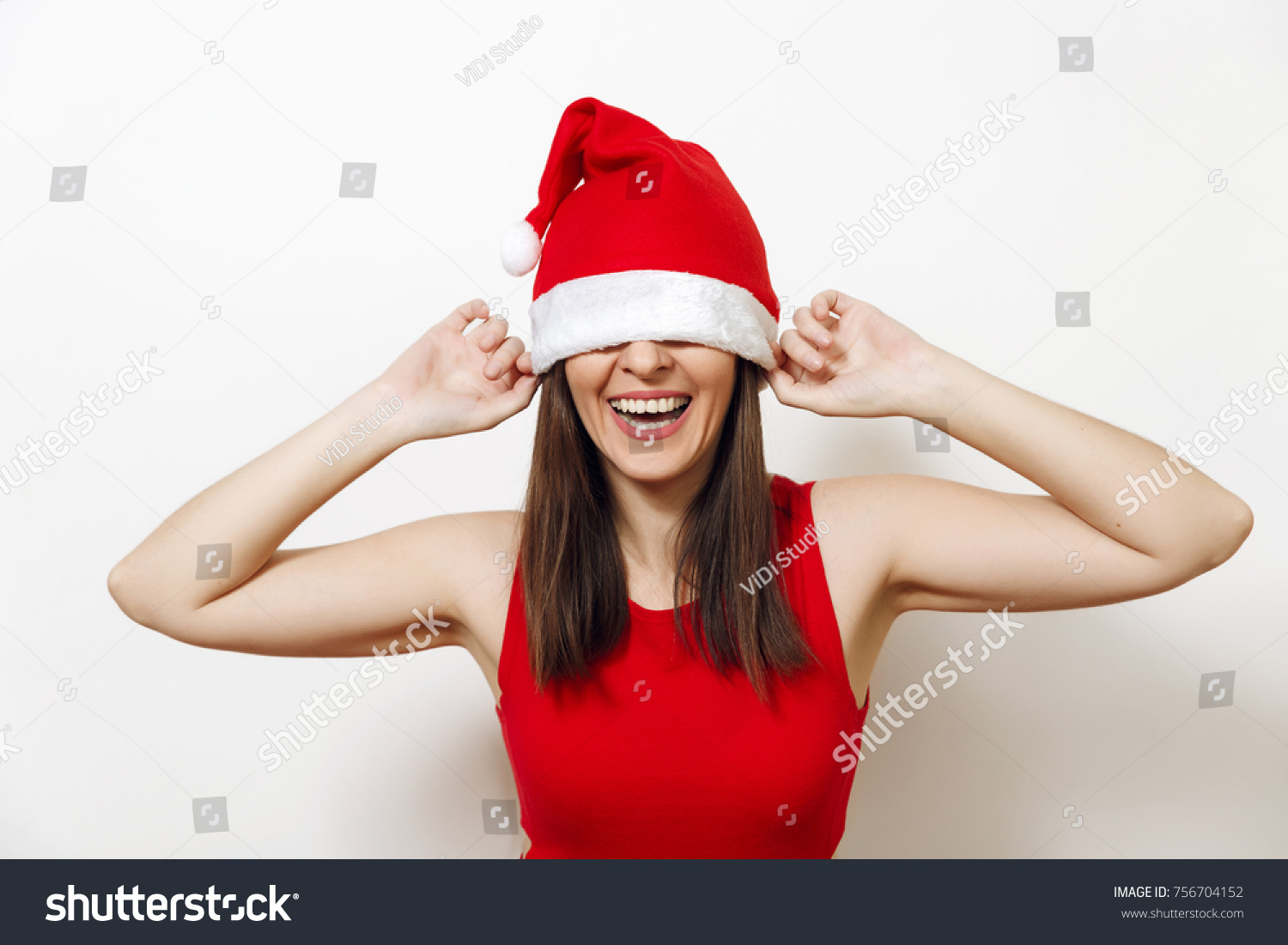 Pretty caucasian young happy woman with healthy skin and charming smile wearing red dress and Christmas hat covering her eyes on white background. Santa girl isolated. New Year holiday 2018 concept #756704152