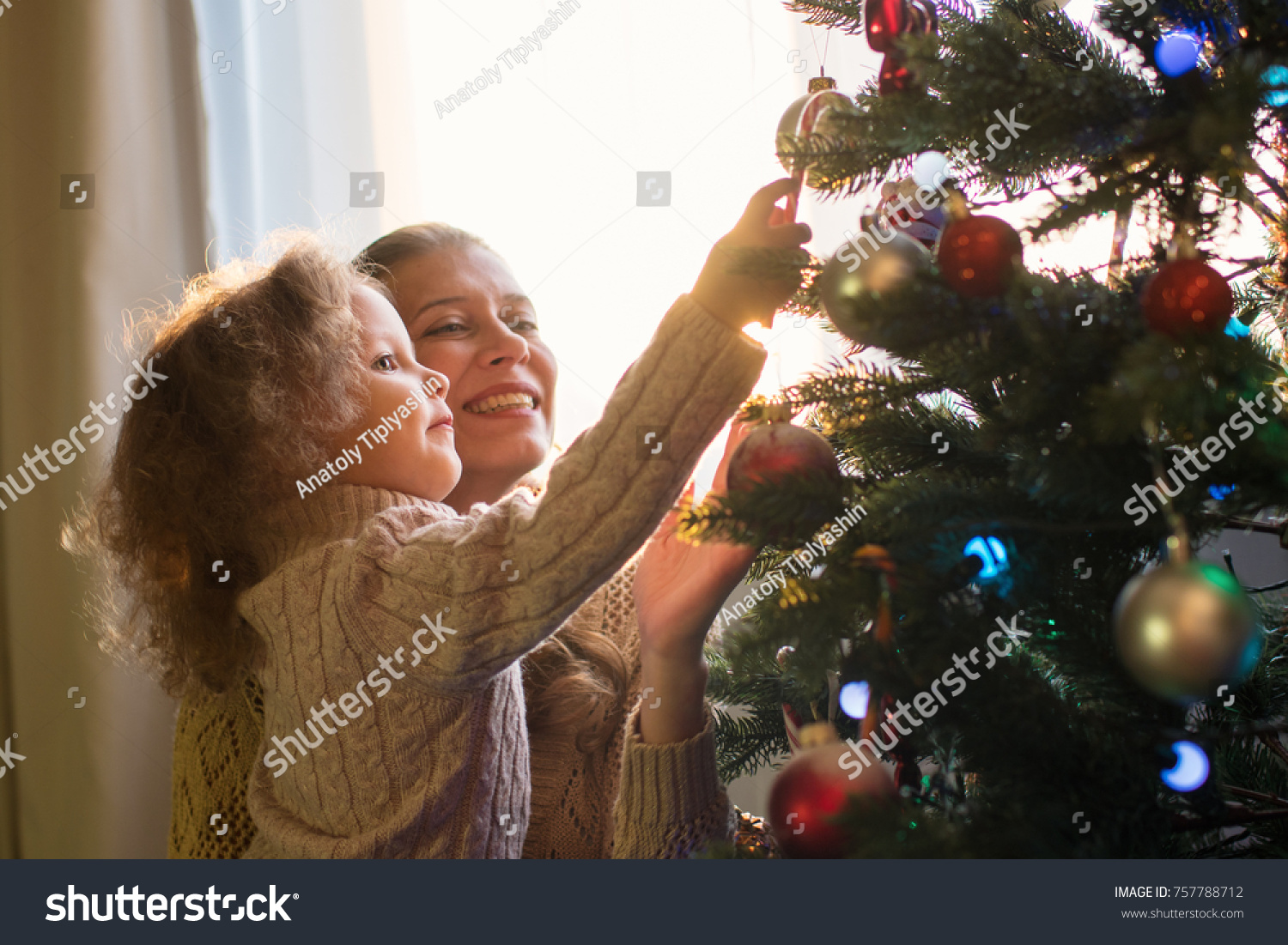 Mother and daughter decorate a Christmas tree against the window with a setting sun and bright sunlight #757788712