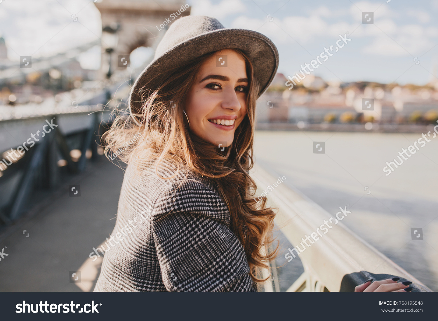Outdoor photo of romantic european woman with curly hairstyle spending time outdoor, exploring european city. Graceful young lady in gray coat and hat enjoying views on embankment. #758195548